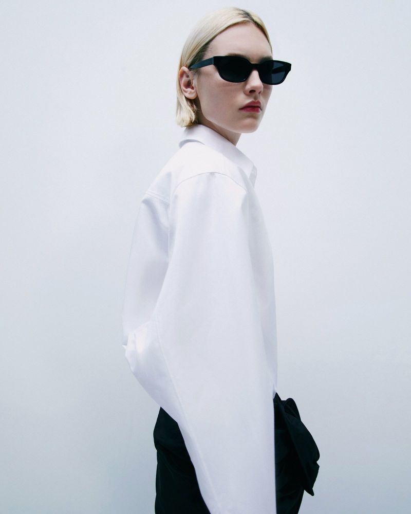 A young adult woman with platinum hair in a white button-down and black bottoms poses indoors with a white background wearing sunglasses.