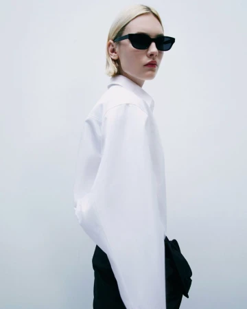 A young adult woman with platinum hair in a white button-down and black bottoms poses indoors with a white background wearing sunglasses.