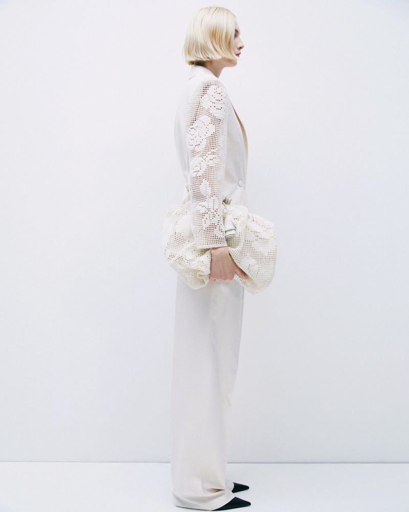 Blond woman in cream-colored wide-leg suit with lace sleeves and large clutch poses in a minimalist studio against a white backdrop.