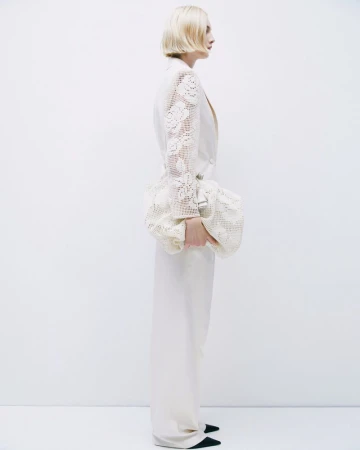 Blond woman in cream-colored wide-leg suit with lace sleeves and large clutch poses in a minimalist studio against a white backdrop.