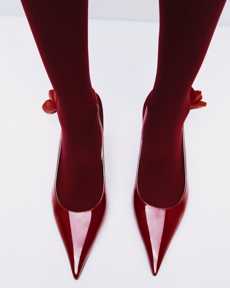 A pair of glossy red patent leather pointed-toe high heels with matching red tights are isolated on a white background.