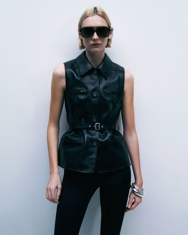 Blonde fashion model in dark sunglasses and belted leather vest stands against white wall. Bracelet and black trousers complete the look.