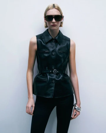 Blonde fashion model in dark sunglasses and belted leather vest stands against white wall. Bracelet and black trousers complete the look.