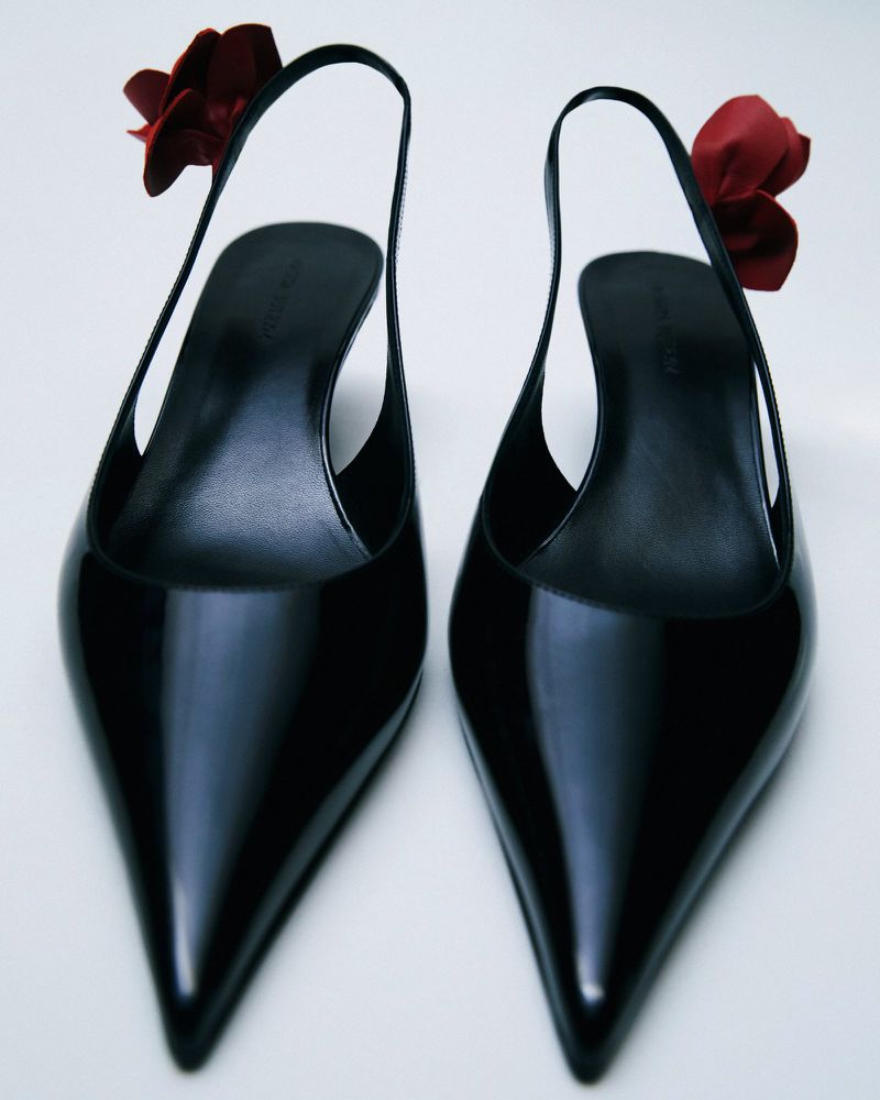 Pointed-toe slingback shoes in black patent leather with red floral embellishments against a white background in a studio shot.