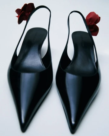 Pointed-toe slingback shoes in black patent leather with red floral embellishments against a white background in a studio shot.