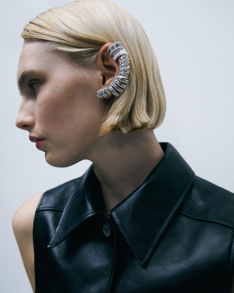 Profile portrait of a blonde female wearing a black collared shirt and a sculptural, crystal ear cuff on a white background.