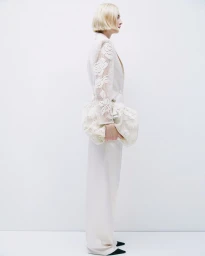 Model with blonde bob wearing an off-white lace sleeve blazer and trousers holding a matching pouch bag against a white seamless background