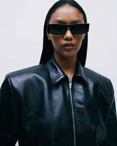 Model in black sunglasses and a black leather zip jacket against a white background for Magda Butrym