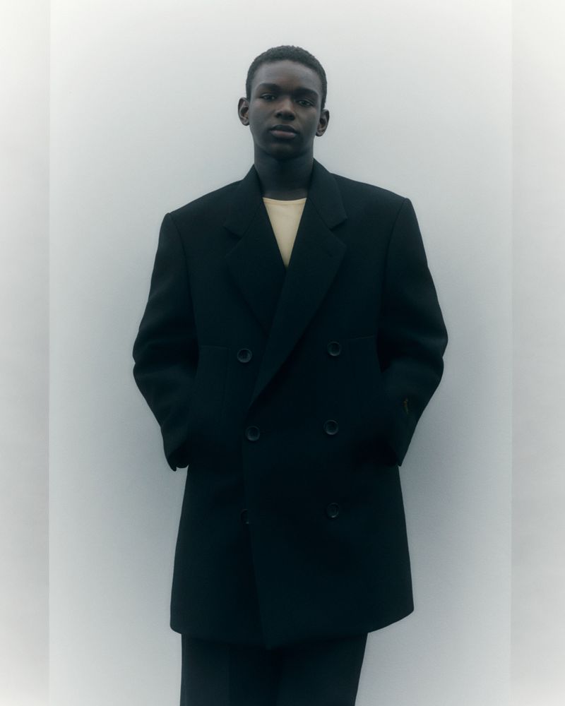 An adult male of African descent poses in a double-breasted black overcoat against a neutral background, showcasing a fashion-forward aesthetic.