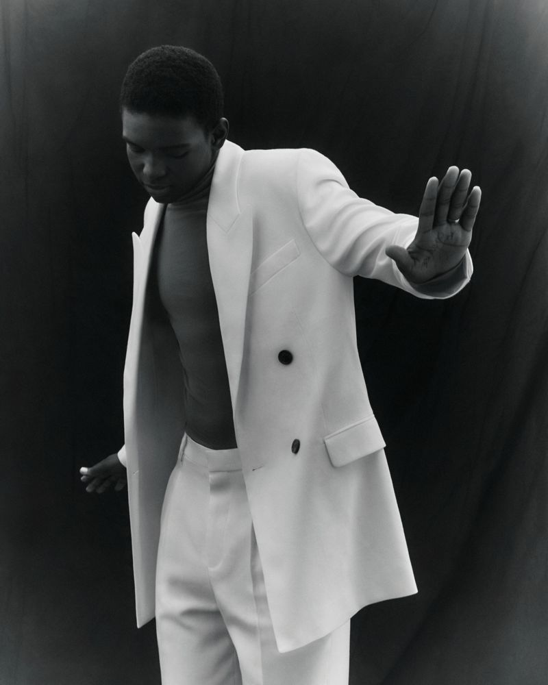 Monochrome portrait of an African American man in a white suit, posing with an open jacket and outstretched arm against a black backdrop.