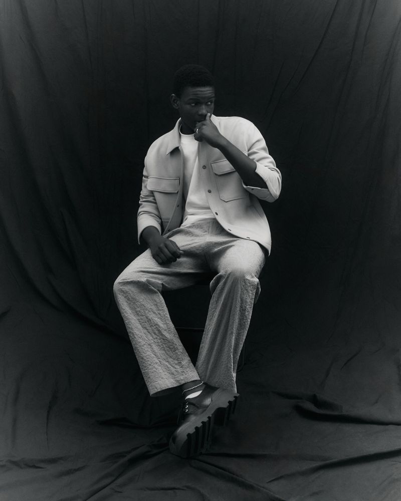 Portrait of a stylish African male model in a light jacket and trousers, seated against a dark backdrop in a studio setting.
