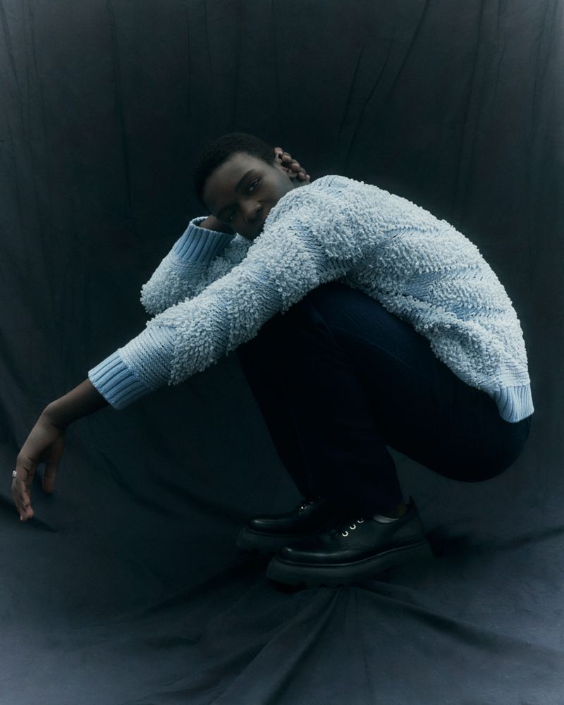 A single young adult poses in a studio wearing a light blue textured sweater, dark trousers, and chunky black shoes, against a gray backdrop.