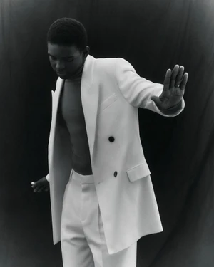 Black man wearing a white double-breasted blazer and trousers raising his hand in a studio portrait for Vogue Italia