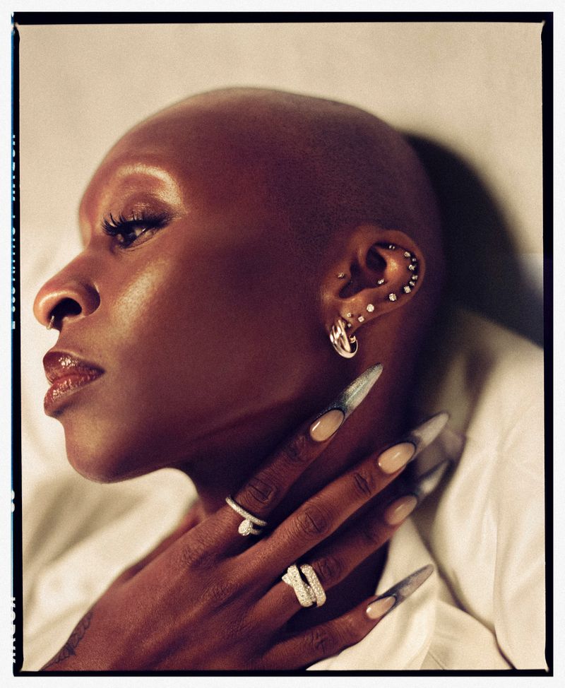 Elegant portrait of an African American woman with a shaved head, statement jewelry, long nails, and a neutral expression, shot in studio.