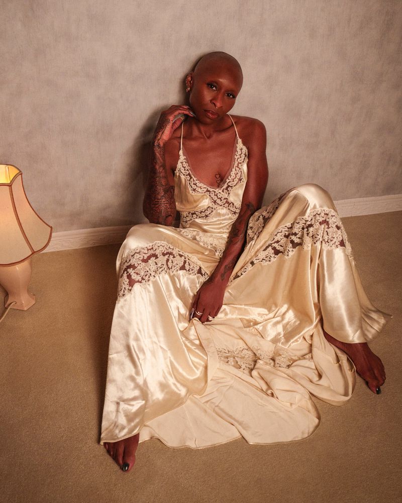 Woman with shaved head poses indoors in a silk dress. She sits on the floor next to a lamp in a dimly lit space.
