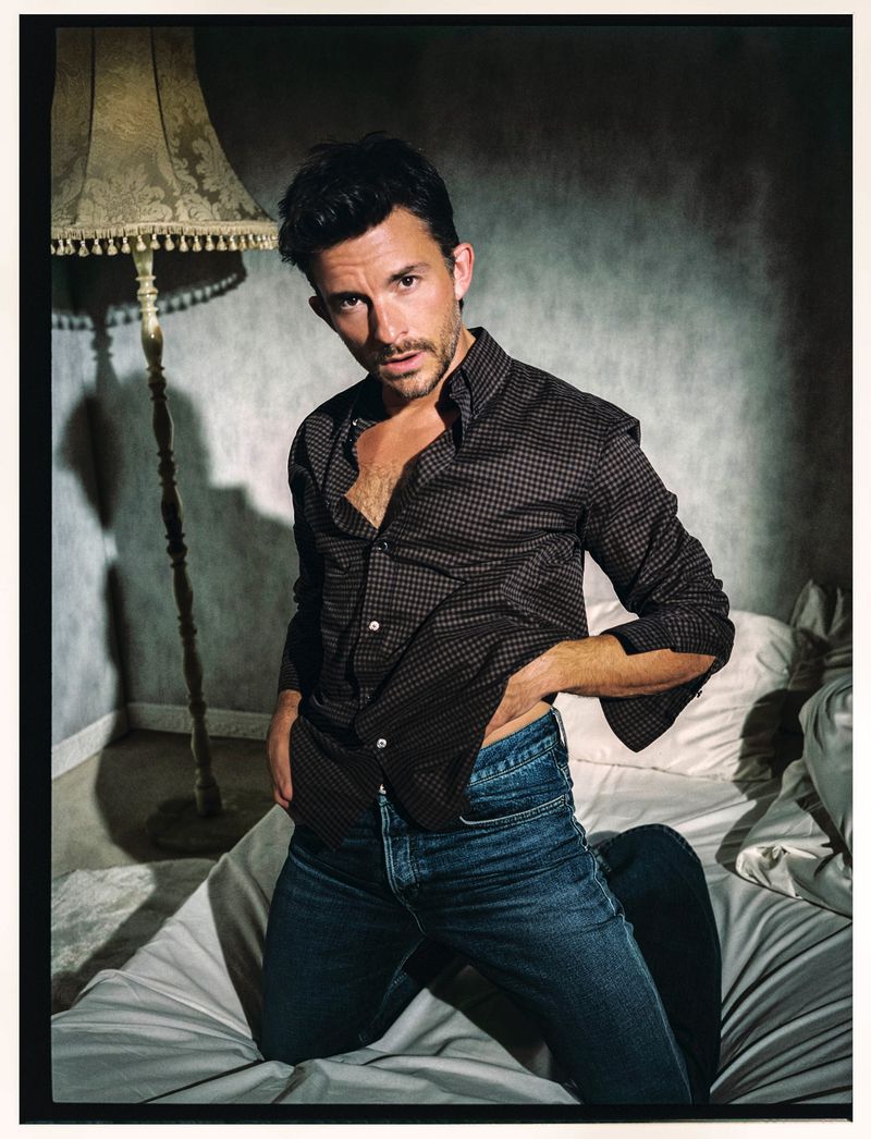 Portrait of a man kneeling on a bed with a patterned shirt, blue jeans, and a lamp in the background, capturing a relaxed and intimate mood.