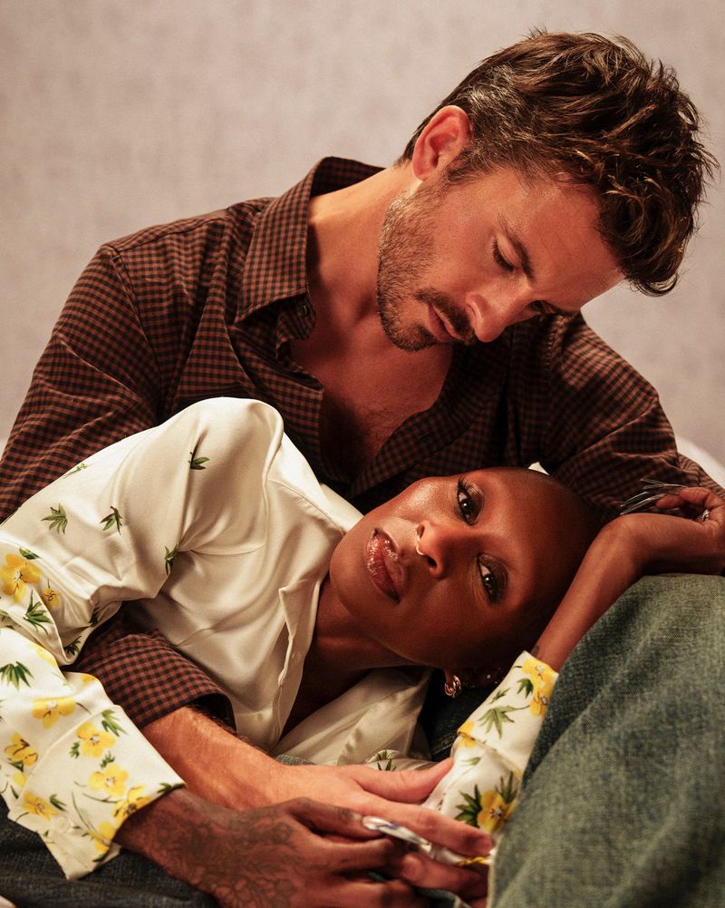 A couple poses intimately in a studio setting. The man with brown hair embraces the bald woman with her head resting on his chest.