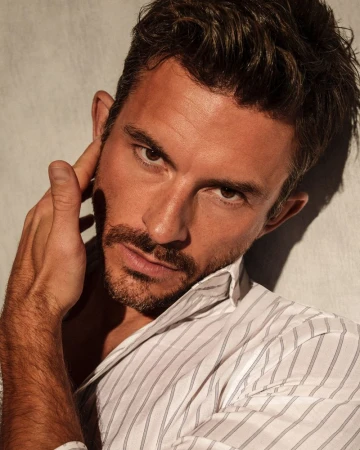 Close-up portrait of an adult man with brown hair and facial hair wearing a white striped button-down shirt, against a neutral background.