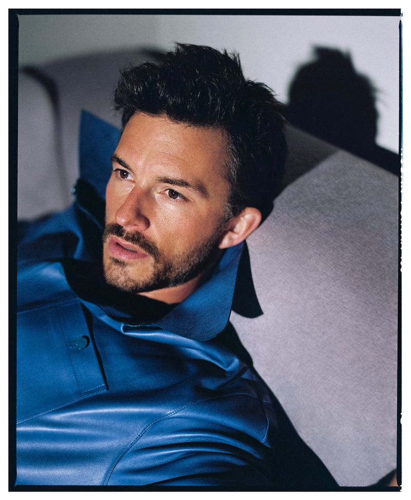 Portrait of a handsome adult male reclining on a sofa in a blue satin jacket. He is lit by natural light in an indoor setting.