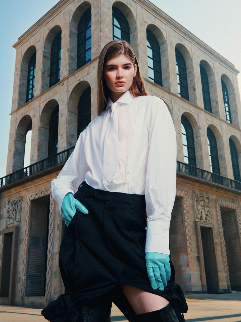 Fashion shot of a young adult woman posing in a white shirt, black skirt, and gloves, in front of a large stone building on a sunny day.