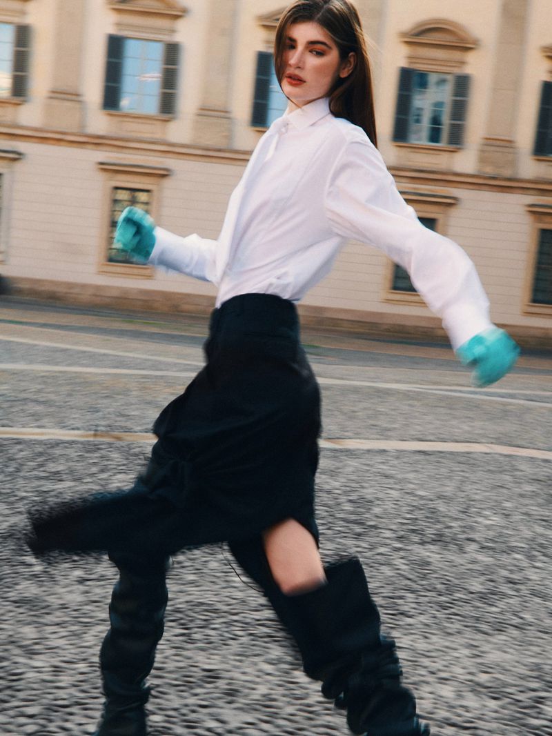 A woman with long brown hair walks in a city square. She wears a white shirt, black skirt, and black boots with aqua gloves. 
