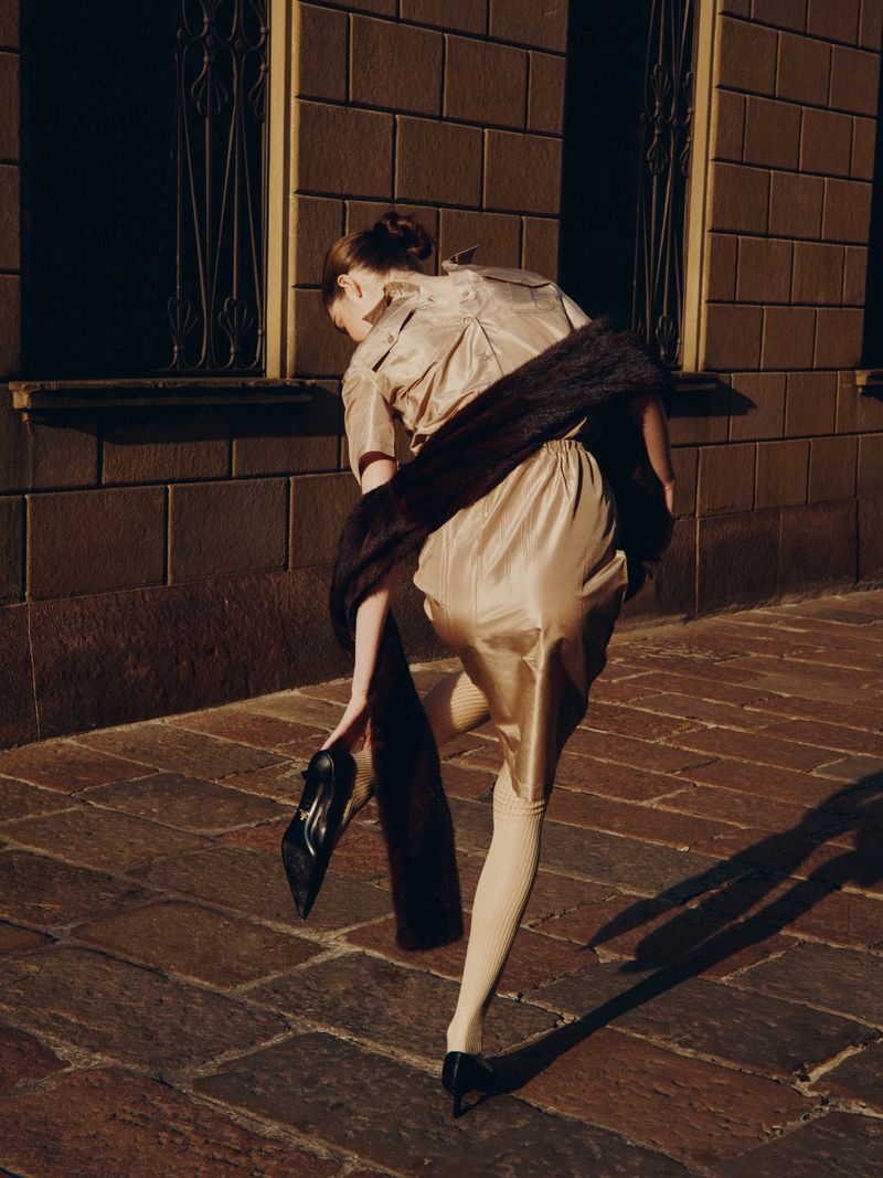 Back view of a woman on a city sidewalk wearing a shiny tan dress and black pumps, holding another pump in her hand.