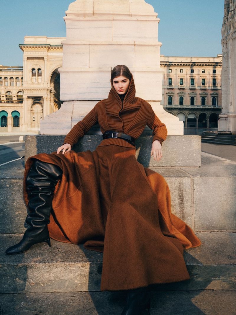 Model seated outdoors in a brown knit hooded sweater and long skirt with black leather boots against classical architecture.