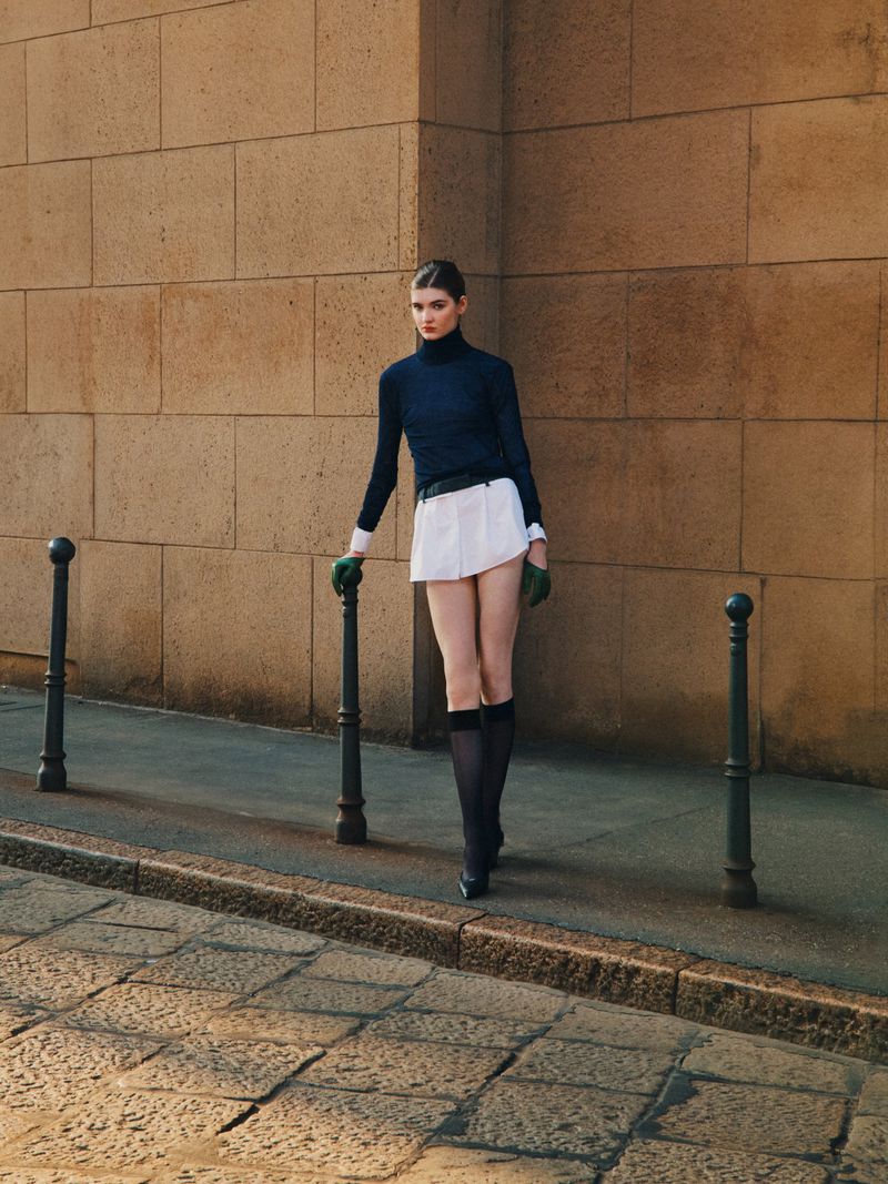 A young woman in a stylish vintage-inspired outfit poses against a stone wall on a city street in bright sunlight, fashion editorial.