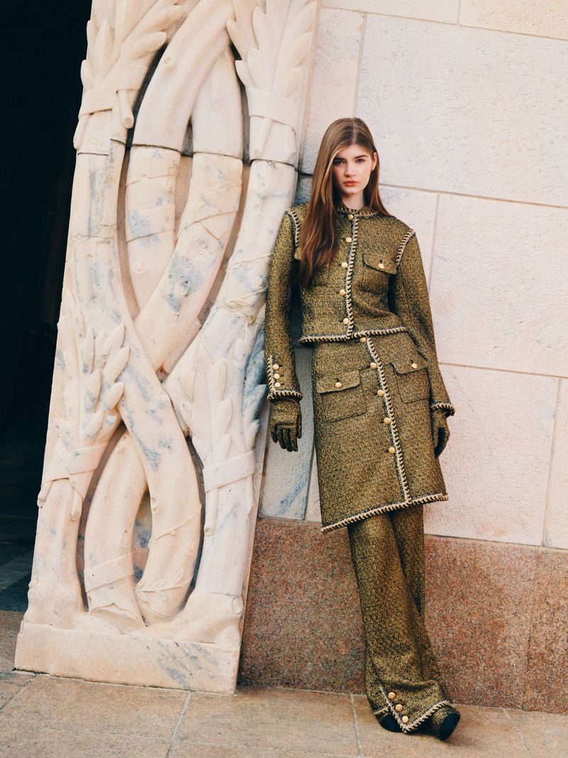 Fashion model in a gold tweed skirt suit leaning against ornate stone architecture for a fashion editorial, outdoors, daytime.