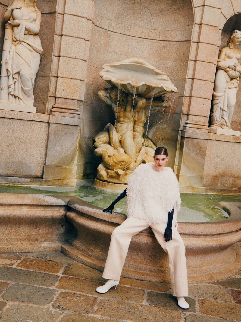 Woman in a white fringed poncho and beige pants poses in front of a stone fountain with statues, shot at eye-level with natural lighting.
