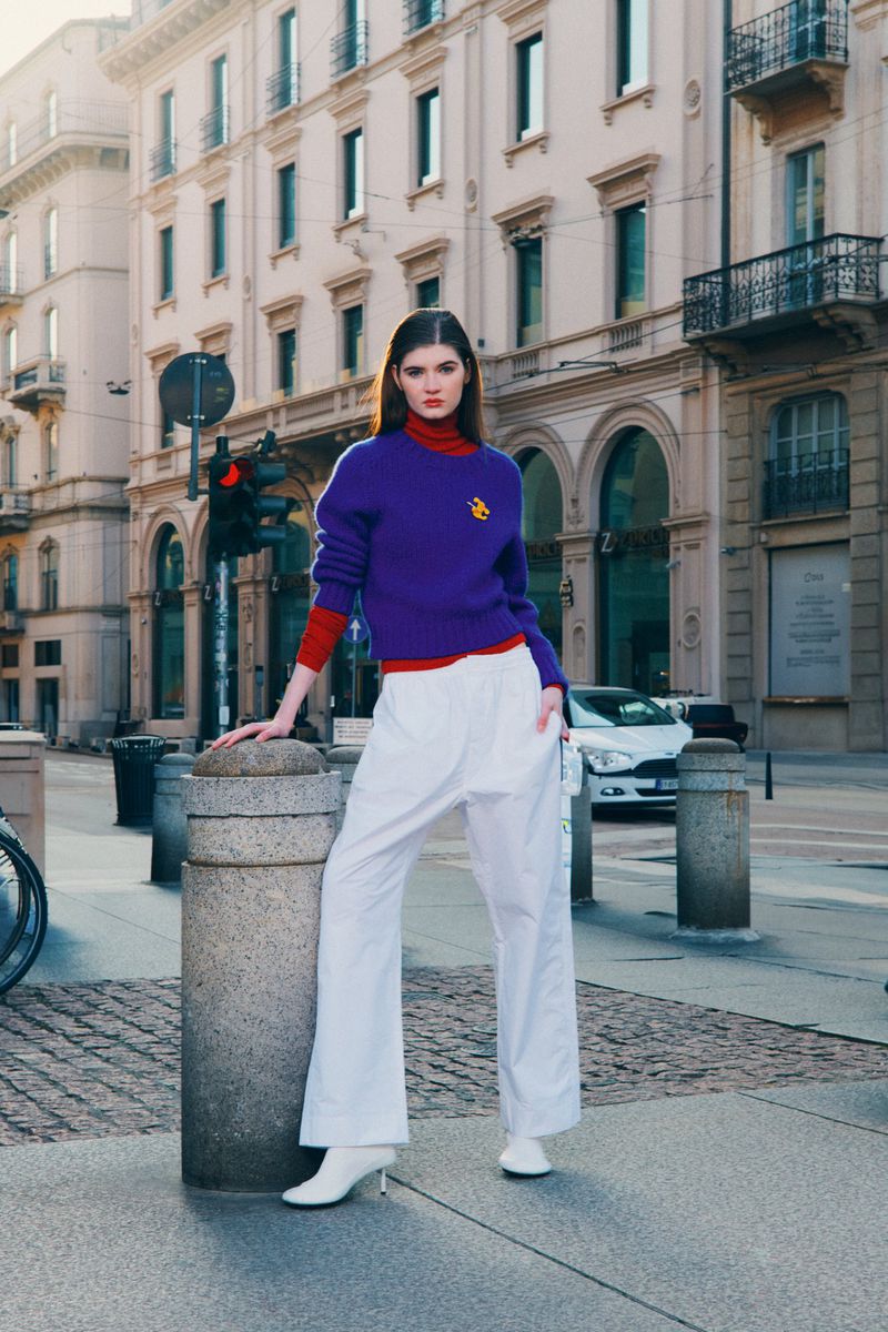 A fashion shot of a young adult woman posing in a purple sweater and white pants against an urban backdrop with classic architecture.