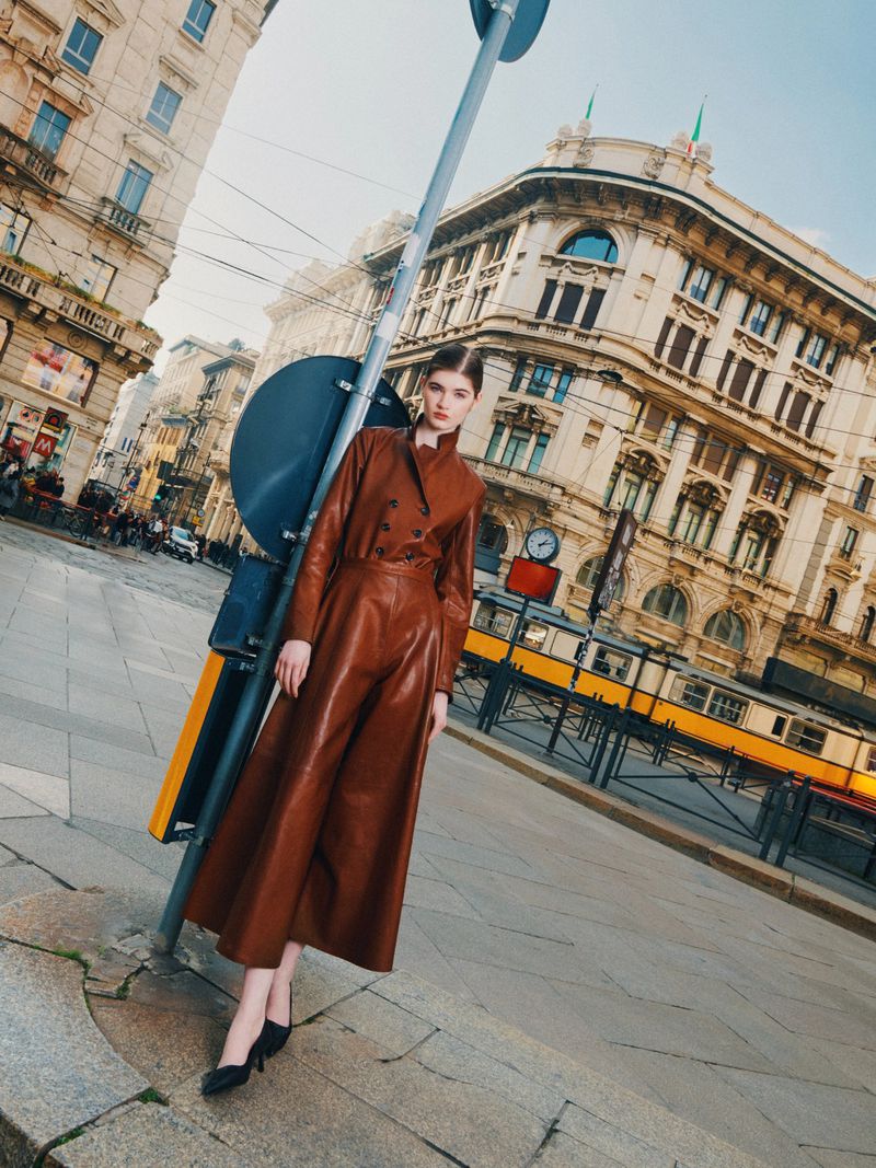 Adult woman in brown leather coat dress stands on a city street next to a traffic signal with buildings and a trolley in the background.