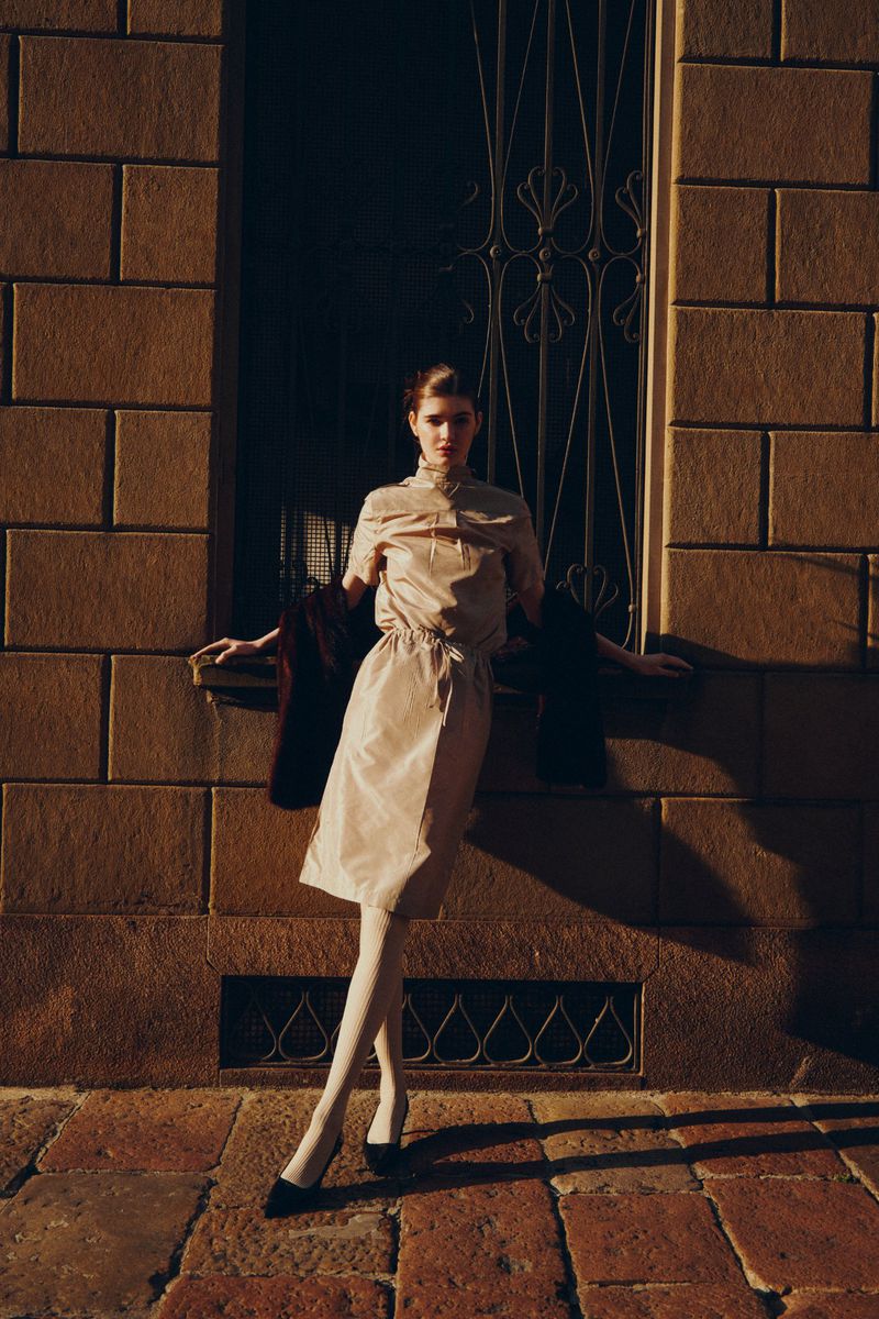 Woman in neutral-toned dress and white tights poses against a brick wall outside with a metal-grate window in soft, diffused sunlight.