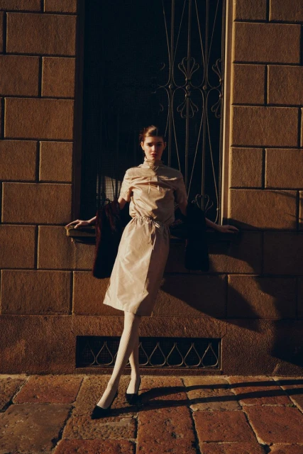 Woman in neutral-toned dress and white tights poses against a brick wall outside with a metal-grate window in soft, diffused sunlight.