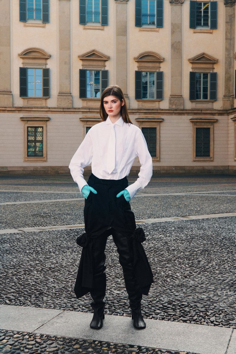 A woman in a white shirt and black pants poses outdoors on a cobblestone surface near a building with many windows and classical facade.
