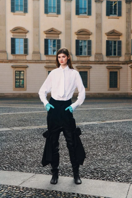 A woman in a white shirt and black pants poses outdoors on a cobblestone surface near a building with many windows and classical facade.