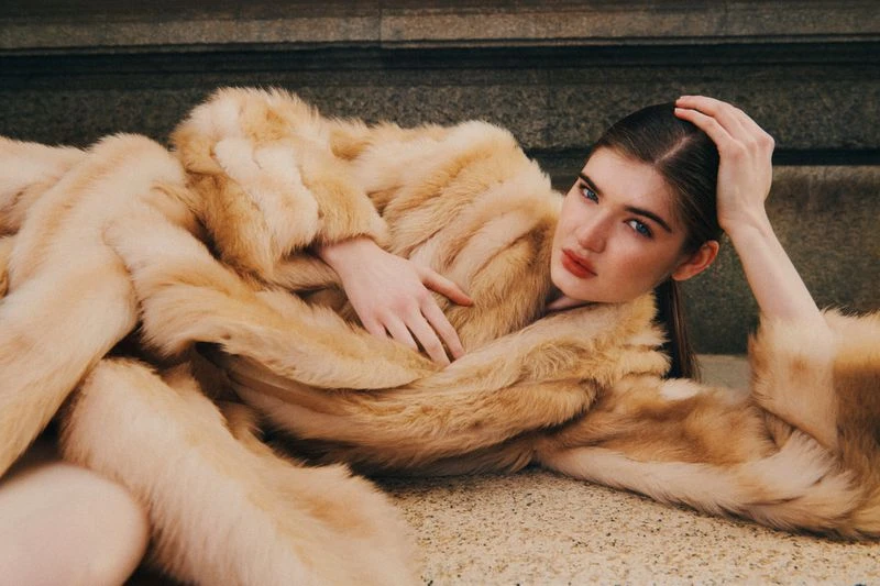 Woman in a fur coat lying down against a gray stone backdrop, posing with her hand on her head and chest, staring at camera.