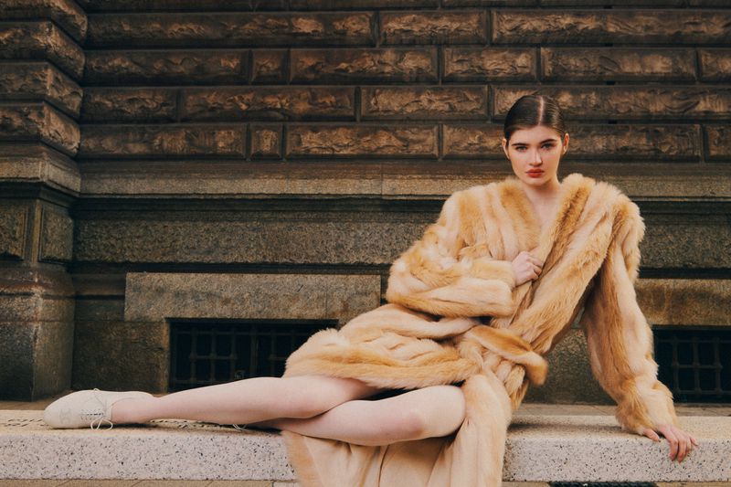 Woman in a fur coat seated on a ledge in front of a brick wall. Stylish outdoor fashion portrait with neutral tones and natural light.