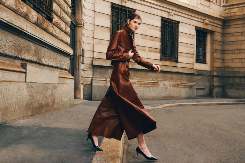 A woman in a brown leather suit walks on a city street with tan stone buildings and black heels; fashion editorial shot with natural light.