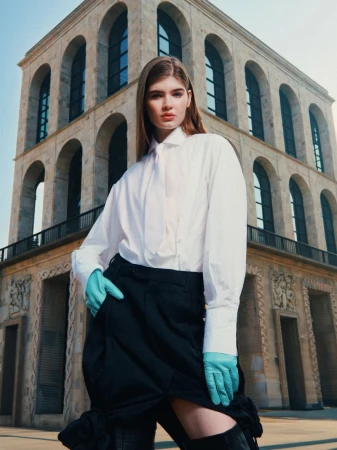 Model wearing a white shirt and black skirt with bright blue gloves posing before a grand classical arched building.