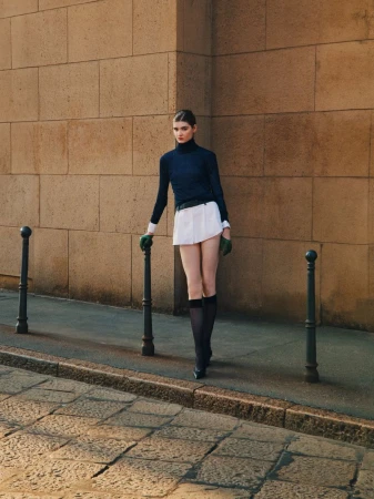 Model standing on a paved street beside a stone wall wearing a dark turtleneck and white skirt
