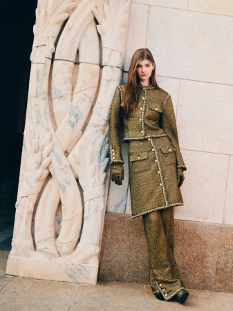Woman wearing an olive green tweed jacket, matching midi skirt, trousers, and gloves leaning against a carved stone wall.