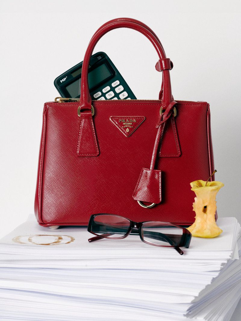 A red Prada handbag sits atop a stack of white papers with glasses, calculator, and apple core on a white background.