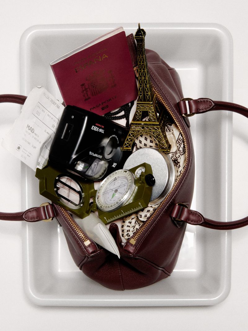 Overhead studio shot of a burgundy leather purse in a white tray, filled with a passport, travel tickets, camera, compass, and Eiffel Tower.