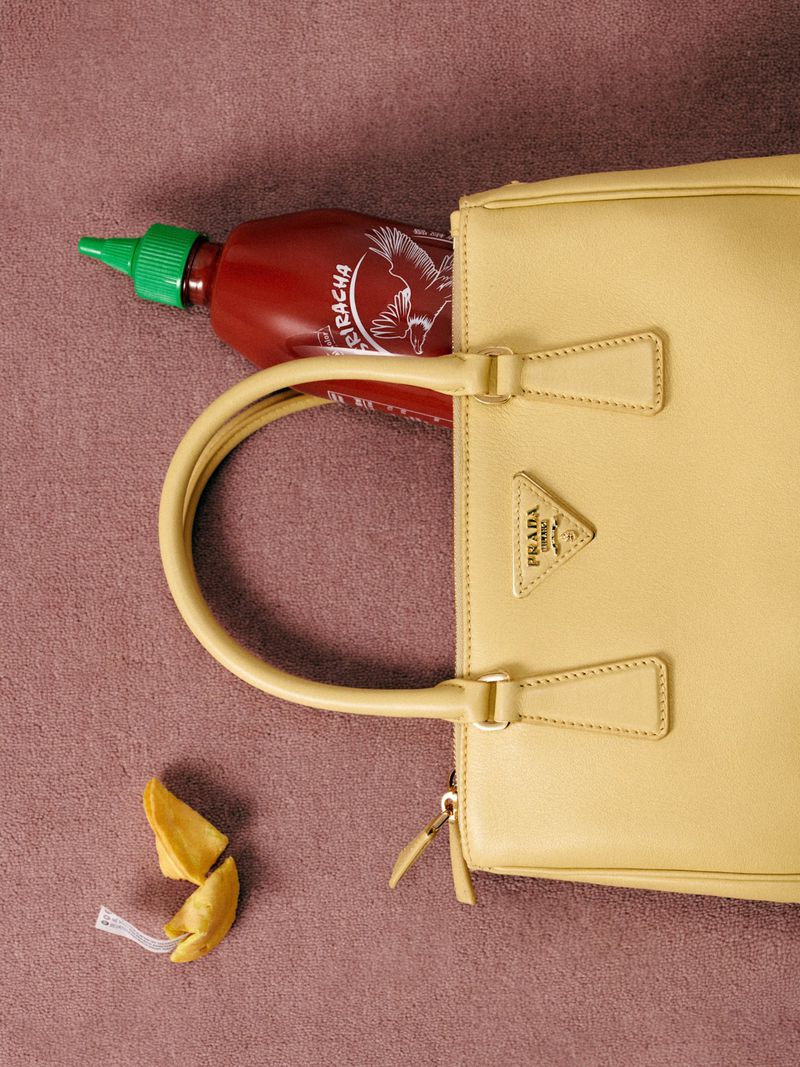 Still life shot of a beige Prada purse with a bottle of Sriracha hot sauce inside and a fortune cookie next to it on a mauve surface.