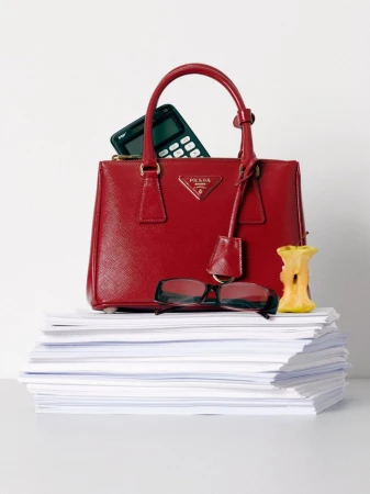 Red Prada handbag sits atop a stack of white paper with eyeglasses and an apple core on a white table against a white background.