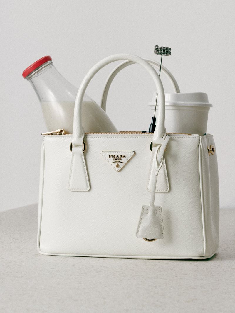 Cream-colored Prada handbag holding a bottle of milk, a beverage cup, and a whisk against a neutral background in a studio setting.