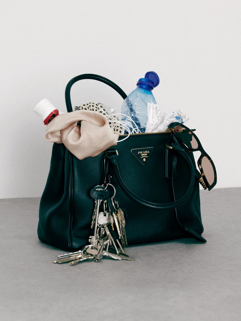Prada handbag overflowing with personal items like keys, water bottle, scrunchie, and sunglasses set against a minimalist gray backdrop.
