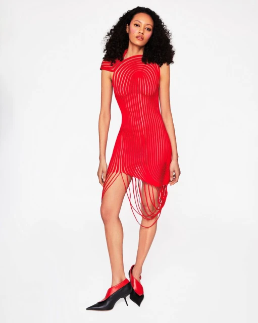 Full-length shot of an adult female fashion model posing in a short, red form-fitting dress with string fringe and black and red heeled shoes.