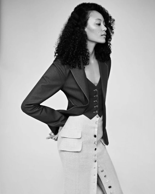 Black and white studio shot of a stylishly dressed, dark-haired woman in a blazer, vest, and skirt, posing with hands on her hips.